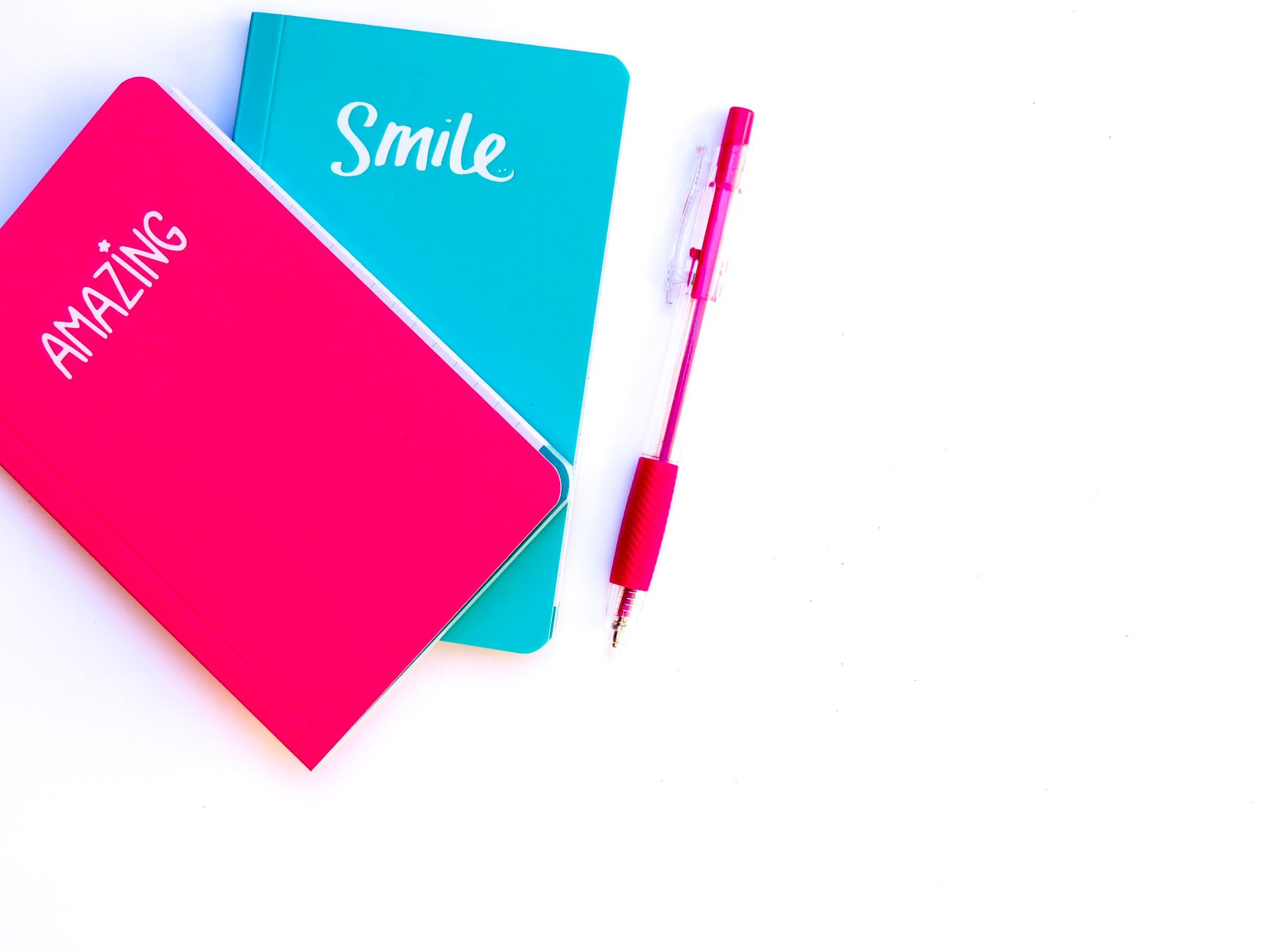 Kleurrijke flatlay met turquoise en roze notitieboeken en bijpassende pen op een witte achtergrond.