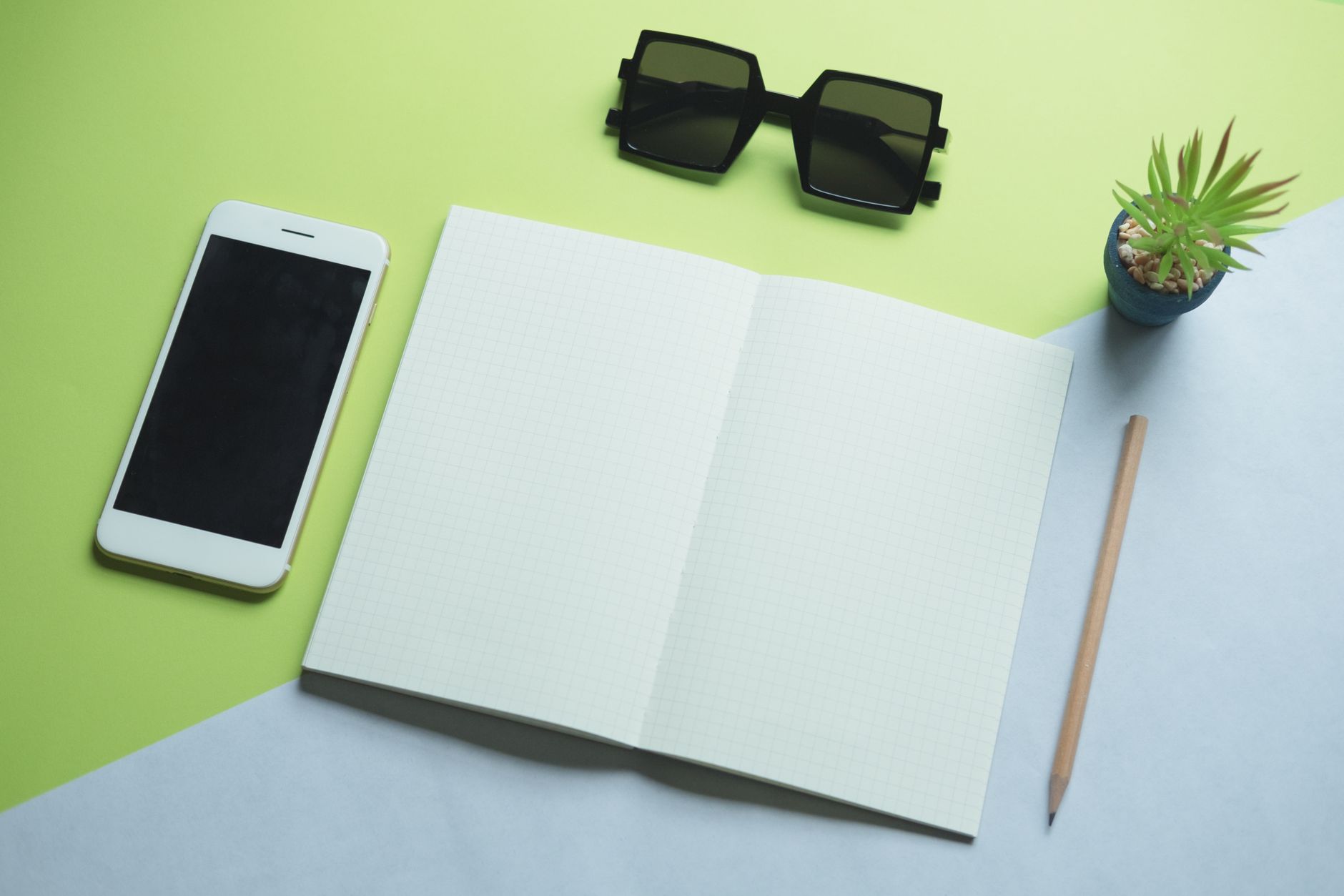 Flatlay van moderne bureau-essentials: smartphone, notitieblok, zonnebril en plant.