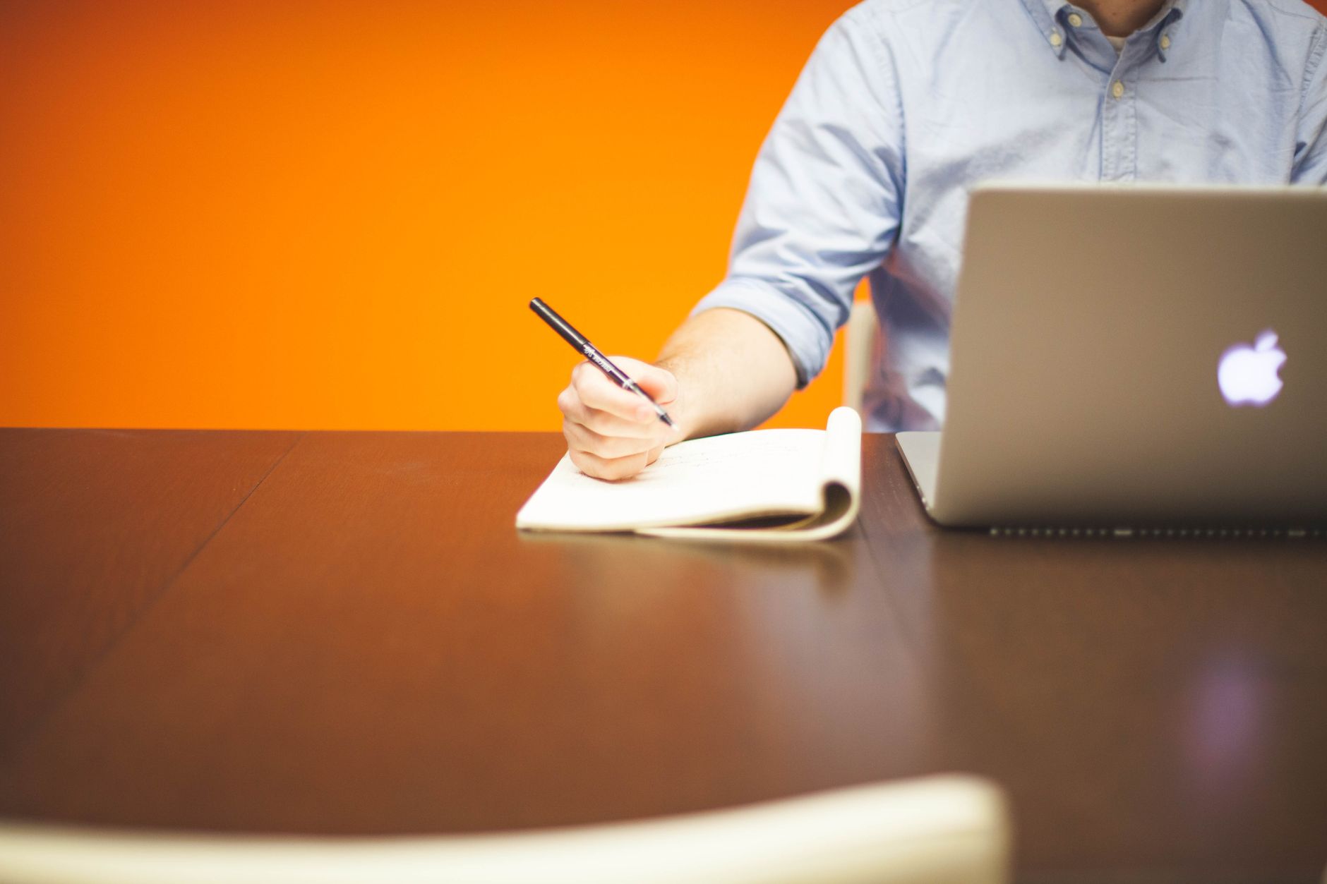 Iemand schrijft notities in een notitieboek naast een laptop op een houten bureau met oranje achtergrond.